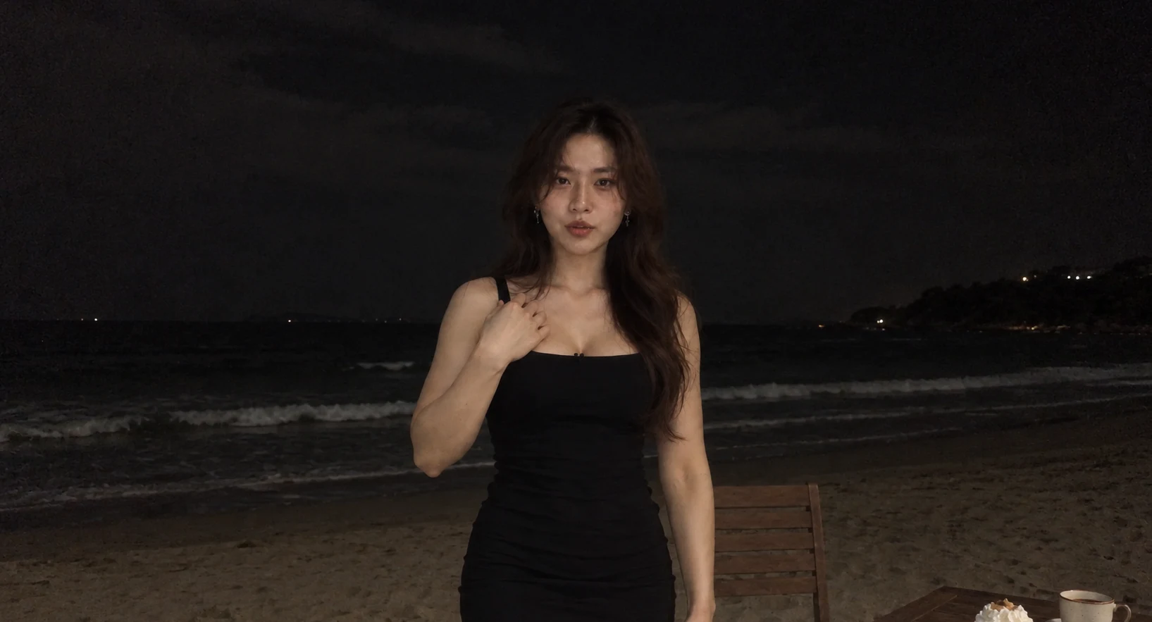 Adult woman in a black dress on a dark beach, with a wooden chair and table beside her.