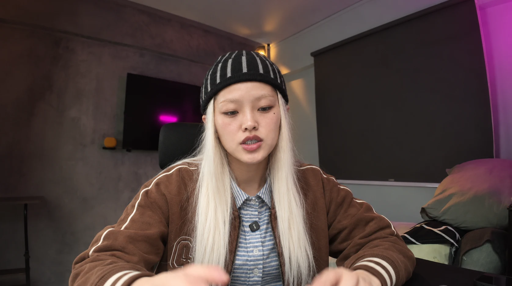 Platinum-haired adult woman in a striped beanie and brown jacket, seated in the original room.